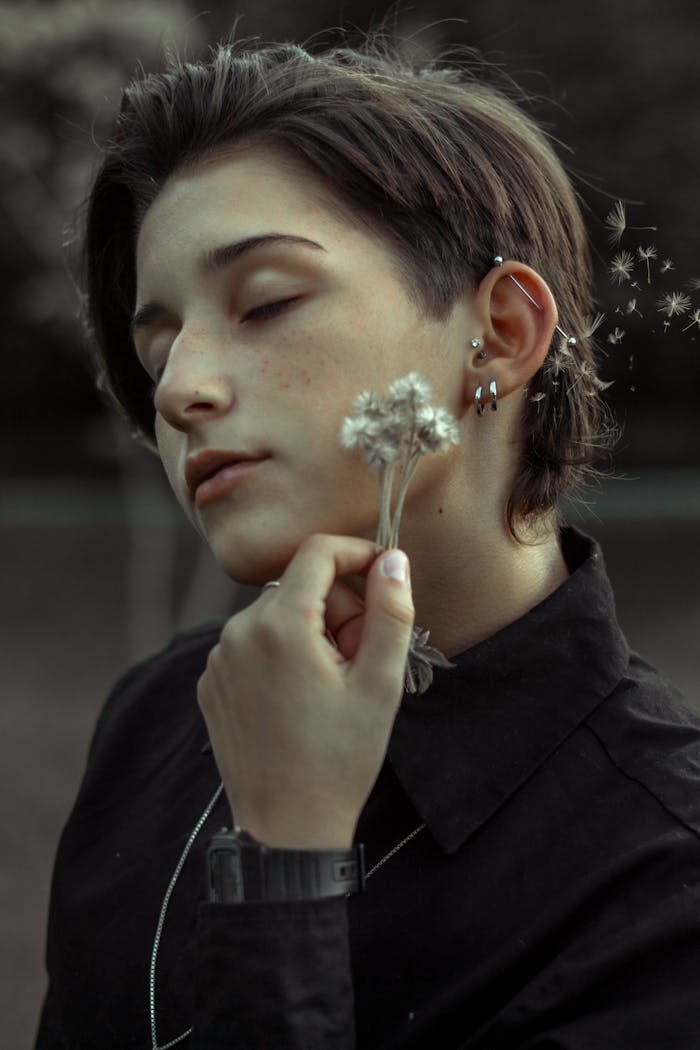 A serene portrait of a young adult with closed eyes, holding dandelions, showcasing piercings and short hair.